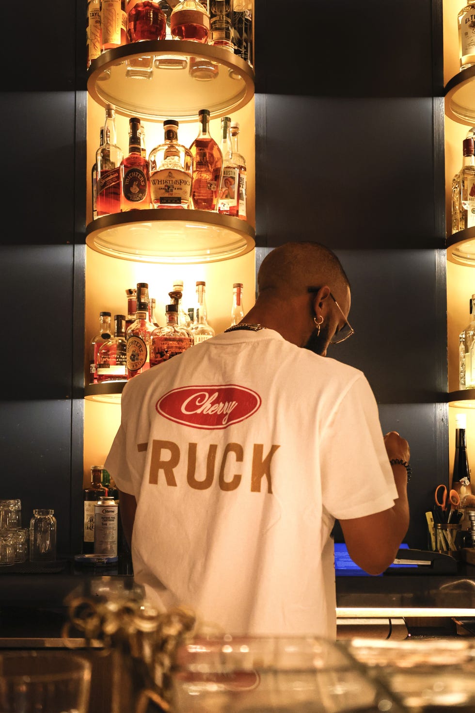 a person in a bar wearing a cherry los angeles shirt and preparing drinks with a selection of spirits in the background