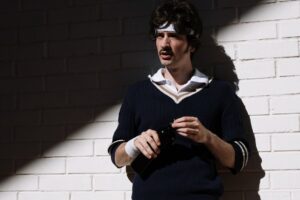 Polo Club Cricket Sweaters Are Creeping Into Off-Duty Tailoring A man wearing a cream cable-knit V-neck sweater layered over a collared shirt with tailored trousers
