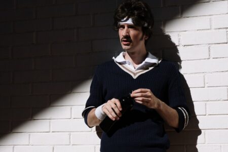 A man wearing a cream cable-knit V-neck sweater layered over a collared shirt with tailored trousers